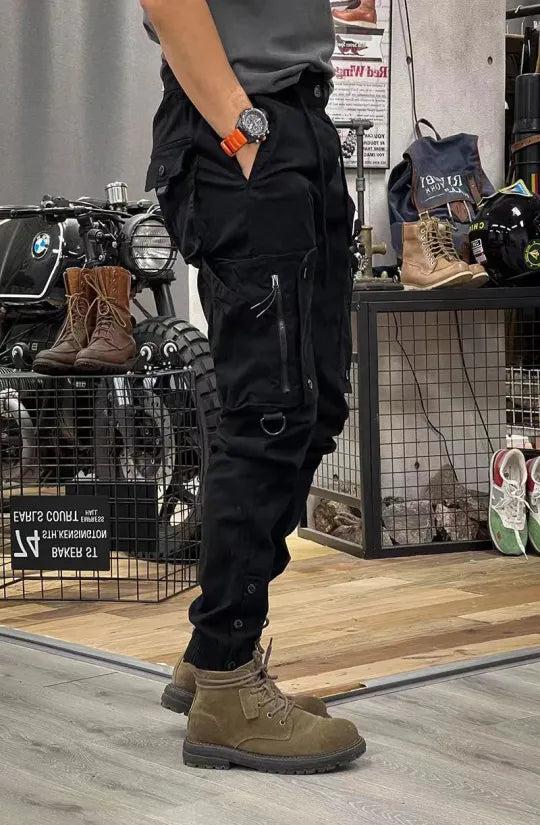 Man in zwarte cargobroek en bruine laarzen, staand naast motorfiets in winkel. Stijlvolle herenkleding, urban mode, motoraccessoires.