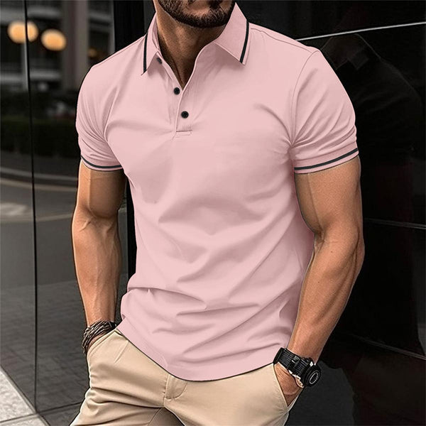Man in roze poloshirt met korte mouwen en beige broek, leunend tegen een muur. Modieuze herenkleding, casual stijl, stedelijke achtergrond.