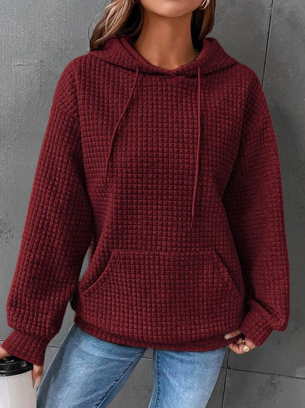 Vrouw in bordeauxrode gebreide hoodie met capuchon en buidelzak, casual stijl, gecombineerd met blauwe jeans, tegen grijze achtergrond.