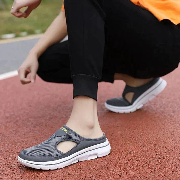 Grijze ademende sportschoenen met witte zolen, gedragen door een persoon in zwarte broek op een rode atletiekbaan. Comfortabele slip-on sneakers.