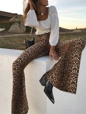 Vrouw in modieuze luipaardprint broek en witte blouse, zittend op een witte muur. Trendy outfit, herfstmode, stijlvolle schoenen, buitenomgeving.