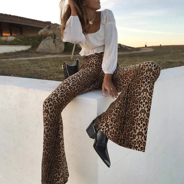 Vrouw in modieuze luipaardprint broek en witte blouse, zittend op een witte muur. Trendy outfit, herfstmode, stijlvolle schoenen, buitenomgeving.