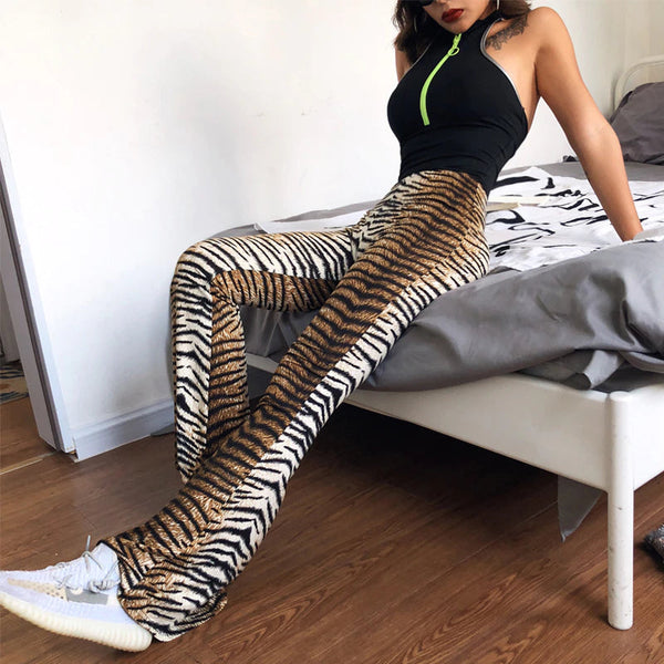 Vrouw in tijgerprint broek en zwarte top zittend op bed, modieuze outfit, trendy kleding, comfortabele stijl, houten vloer, moderne slaapkamer.
