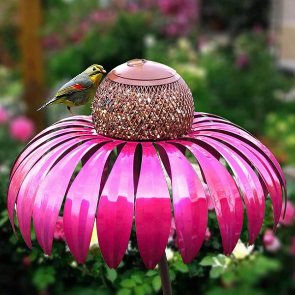 Roze metalen vogelvoeder in bloemvorm met een kleurrijke vogel, omringd door groene tuinplanten. Perfect voor tuinliefhebbers en vogelliefhebbers.