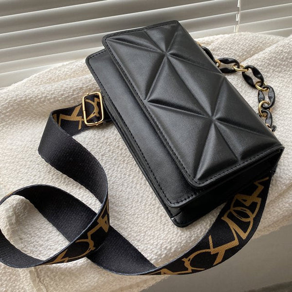 Zwarte leren schoudertas met geometrisch patroon en verstelbare riem met gouden details, liggend op een witte achtergrond bij jaloezieën.
