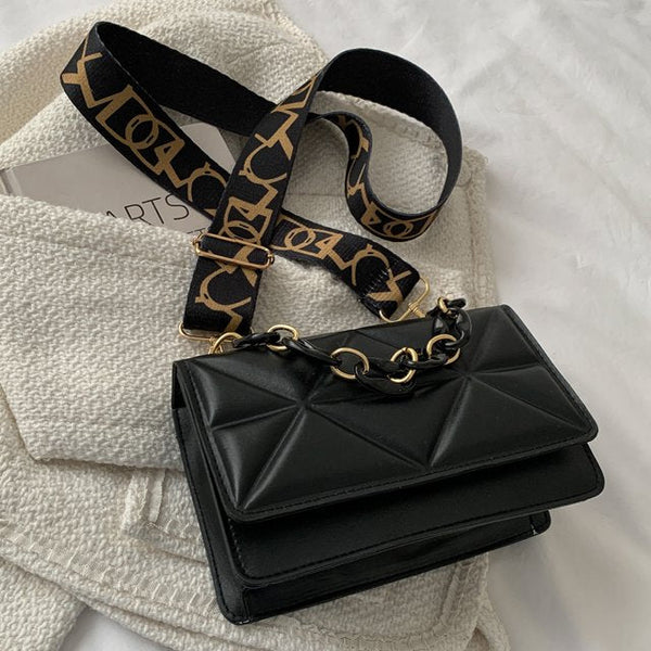 Zwarte leren handtas met geometrisch patroon en goudkleurige kettingriem, liggend op witte stoffen achtergrond. Luxe modeaccessoire.