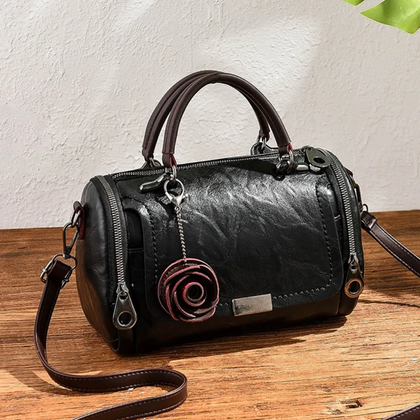 Zwarte leren handtas met rozenhanger, dubbele ritsen en verstelbare schouderriem op houten tafel. Stijlvolle damesaccessoire.