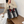 Vrouw met zwarte en witte gestreepte handtas, zittend op houten tafel, draagt beige trui en witte broek, houdt koffiebeker vast.