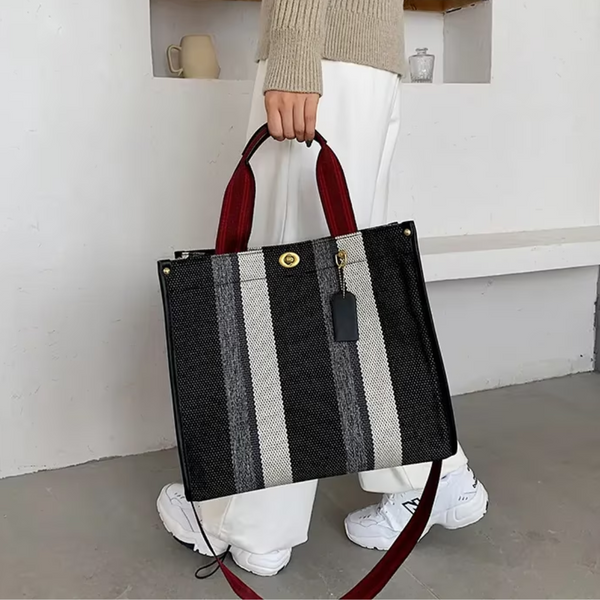 Zwart-witte gestreepte handtas met rode handvatten, gedragen door persoon in witte broek en beige trui. Modieuze tas, casual stijl, trendy accessoire.