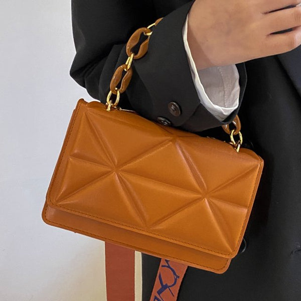 Bruine leren handtas met geometrisch patroon en gouden kettingriem, gedragen door persoon in zwarte jas. Stijlvolle damesmodeaccessoire.