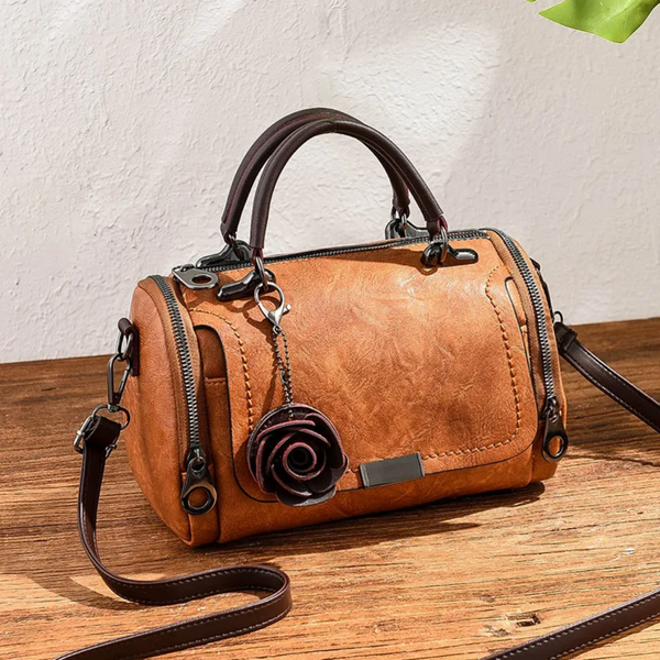 Bruine leren handtas met rozenhanger, cilindervormig ontwerp, dubbele ritsen en verstelbare schouderriem, perfect voor damesmode en accessoires.
