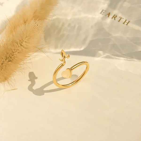Gouden ring met hart- en bloemmotief op beige achtergrond, naast gedroogd gras. Elegante sieraden, minimalistisch design, romantisch cadeau-idee.