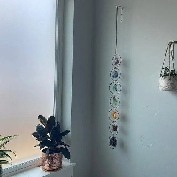 Chakra wanddecoratie met zeven edelstenen, hangend aan een muur naast een raam. Planten in koperen potten op de vensterbank.