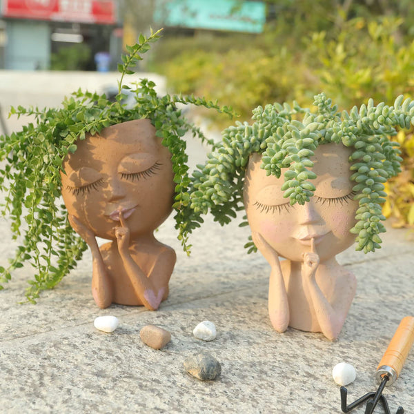 Twee creatieve plantenpotten in de vorm van gezichten met groene planten als haar, geplaatst op een stenen oppervlak in een tuinomgeving.