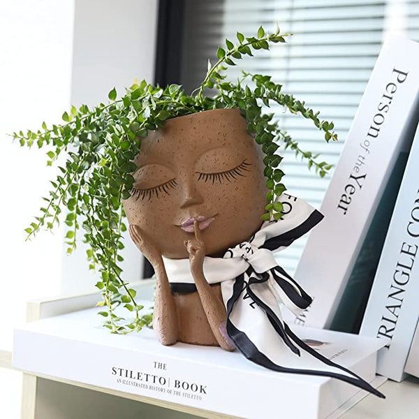 Decoratieve plantenpot in de vorm van een gezicht met gesloten ogen, gevuld met groene hangplant, geplaatst op boeken. Perfect voor moderne interieurdecoratie.