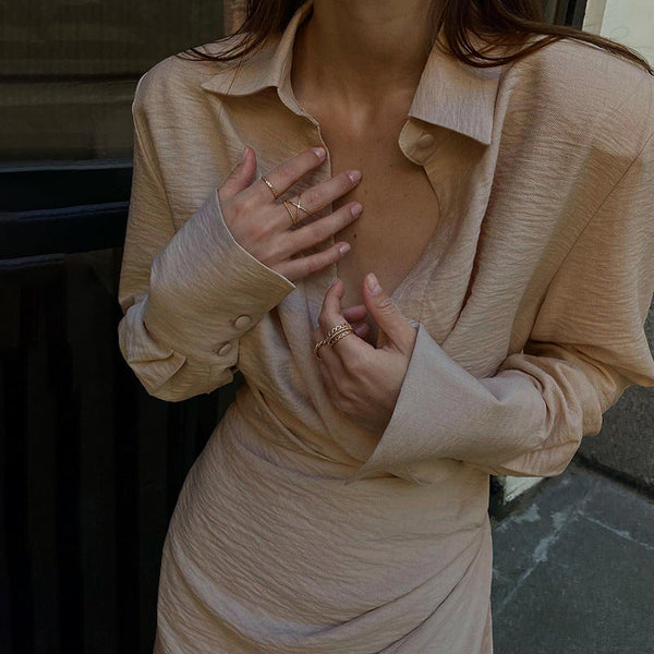 Vrouw in beige blouse en rok, met gouden ringen, poseert voor een donkere achtergrond. Elegante mode, minimalistische stijl, trendy outfit.