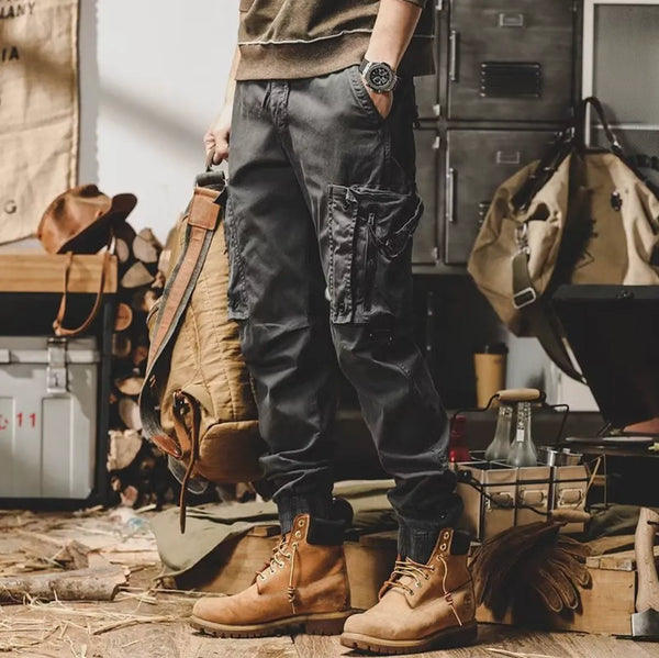 Man in casual outfit met grijze cargobroek en bruine laarzen, staand in een rustieke kamer met houten vloer en vintage decor.