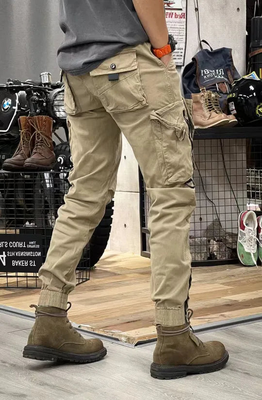 Beige cargobroek met zakken, gedragen door persoon in winkel. Outdoor laarzen en motoraccessoires op achtergrond. Casual mode, herenkleding.