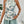 Vrouw in zomerse outfit met haltertop met tropische print en mintgroene shorts, tegen een witte achtergrond met planten. Perfect voor strandmode.