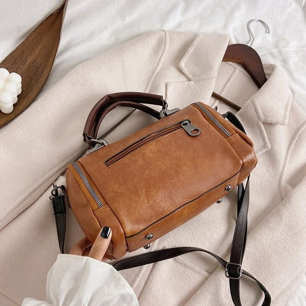 Bruine leren handtas met rits en verstelbare schouderriem op beige jas. Stijlvolle damesaccessoire, perfect voor dagelijks gebruik.