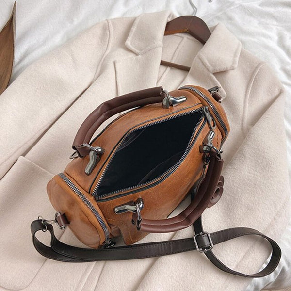 Bruine leren handtas met zilveren ritsen en verstelbare schouderriem, liggend op een crèmekleurige jas. Stijlvolle damesaccessoire.