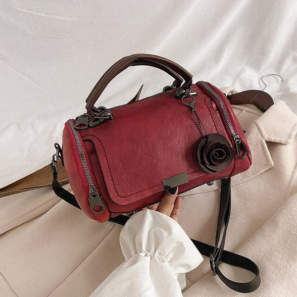 Rode leren handtas met roosdecoratie, dubbele ritsen en verstelbare schouderriem op witte achtergrond. Stijlvolle damesaccessoire.