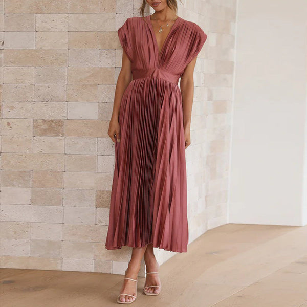 Vrouw in elegante, geplooide, roze jurk met diepe V-hals, staand tegen een stenen muur. Perfect voor avondkleding of formele evenementen.