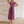 Vrouw in elegante, paarse, geplooide jurk met V-hals, staand tegen een stenen muur. Perfect voor mode, avondkleding, zomerjurken.