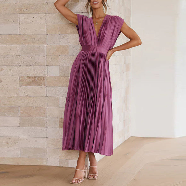 Vrouw in elegante, paarse, geplooide jurk met V-hals, staand tegen een stenen muur. Perfect voor mode, avondkleding, zomerjurken.