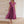 Vrouw in elegante, paarse geplooide jurk met V-hals, staand tegen een beige stenen muur. Perfect voor avondkleding of formele evenementen.