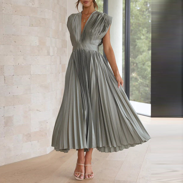 Elegante geplooide grijze jurk, mouwloos met V-hals, gedragen door een vrouw in een moderne interieursetting. Perfect voor formele gelegenheden.