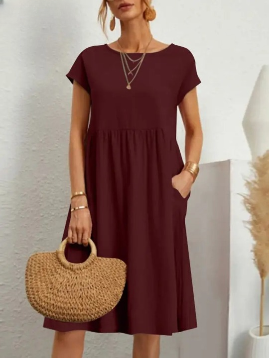 Vrouw in bordeauxrode jurk met korte mouwen, draagt een strooien handtas en gouden sieraden. Stijlvolle zomeroutfit, casual en elegant.