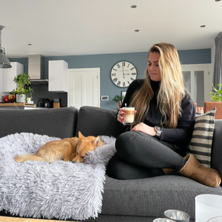 Vrouw zit op grijze bank met slapende Pomsky-hond op een pluizig kleed, moderne woonkamer, koffie in hand, klok aan muur.