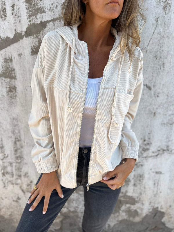 Vrouw in beige corduroy jack met capuchon, witte top en donkere jeans tegen een betonnen muur. Modieuze casual dameskleding, herfststijl.