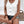 Vrouw in witte mouwloze top met knopen, denim shorts, houten armbanden, zittend voor een witte achtergrond. Zomermode, casual dameskleding.