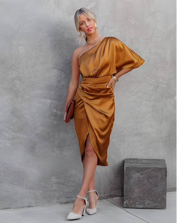 Vrouw in elegante, goudkleurige asymmetrische jurk met clutch en witte hakken, leunt tegen betonnen muur. Mode, avondkleding, stijlvol design.
