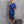Vrouw in elegante blauwe asymmetrische jurk met ruches, staand tegen een grijze muur. Ze draagt een beige handtas en sandalen. Fashion, avondkleding.