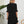 Vrouw in zwarte jurk met ruches, staand voor witte muur met plant en houten stoel. Elegante damesmode, minimalistisch interieur.