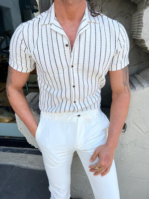 Man in gestreept wit overhemd en witte broek, staand voor een winkel. Modieuze herenkleding, casual zomeroutfit, stijlvolle uitstraling.