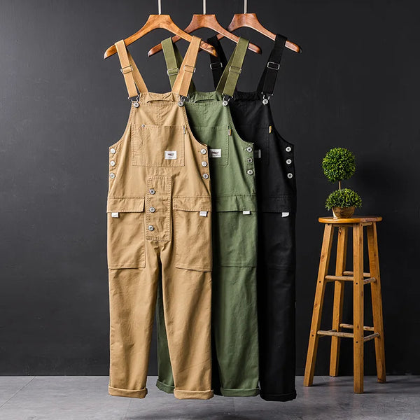 Drie heren tuinbroeken in beige, groen en zwart op houten hangers tegen een donkere achtergrond. Casual zomer jumpsuit met cargozakken.