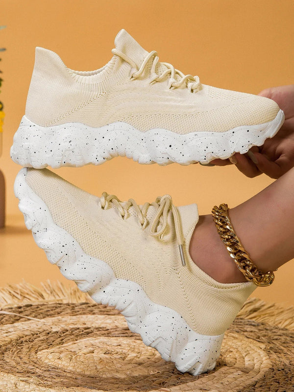 Beige sneakers met dikke, golvende witte zolen en zwarte spikkels, gedragen met een gouden enkelband. Trendy, comfortabele schoenen voor dagelijks gebruik.