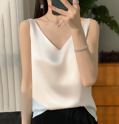 Vrouw in elegante witte satijnen top met V-hals, zilveren sieraden en zwarte broek, gefotografeerd binnenshuis. Stijlvolle mode, minimalistisch design.