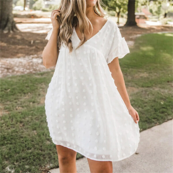 Witte zomerjurk met stippenpatroon, korte mouwen en V-hals, gedragen door vrouw in groene tuin. Perfect voor casual en zomerse outfits.