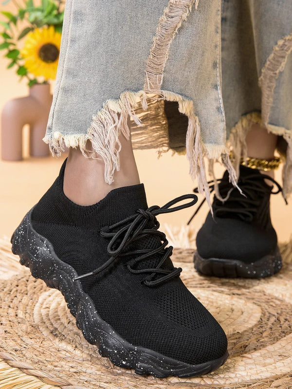 Zwarte gebreide sneakers met dikke zool, gedragen met gerafelde jeans op een rieten mat. Comfortabele damesmode, casual schoenen, trendy stijl.