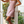 Vrouw in roze bloemenjurk poseert op veranda, omringd door groene planten. Zomermode, casual stijl, elegante sandalen.