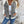 Vrouw draagt een stijlvolle jas met grijze denim accenten en witte mouwen, gecombineerd met een witte top en blauwe jeans, tegen een ruwe muur.
