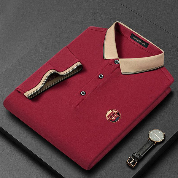 Rode polo met beige kraag, opgevouwen op grijze achtergrond, naast een zwart horloge. Luxe herenkleding, stijlvolle casual mode.