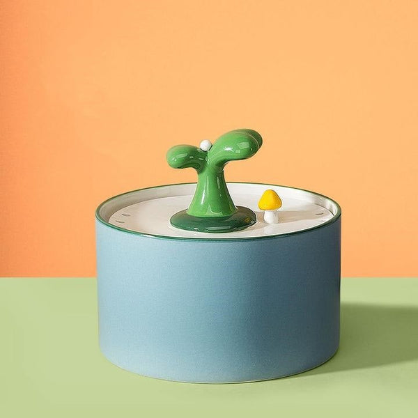Keramische kattenfontein met groen plantontwerp en gele paddenstoel, op een pastelgroene en oranje achtergrond. Stijlvol en functioneel huisdieraccessoire.