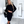 Vrouw in zwarte gebreide jurk met lange mouwen, zwarte laarzen en schoudertas, staand in moderne woonkamer. Elegante mode, herfstkleding.