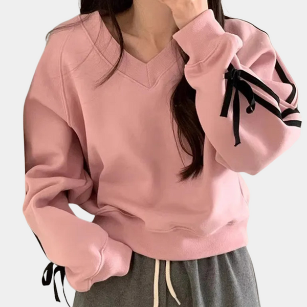 Vrouw in roze oversized trui met zwarte strikdetails op de mouwen, gecombineerd met grijze joggingbroek. Comfortabele casual mode.